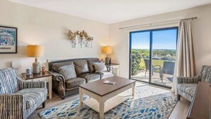 Habitación de hotel con sofá, sillones, una mesita de centro, lámpara, un cuadro y un amplio ventanal corredera que da a un espacio exterior.