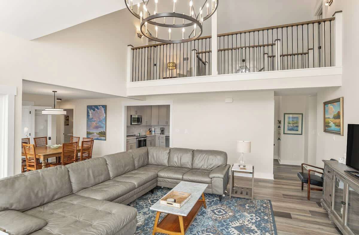 A spacious living area with a large L-shaped sectional sofa, a dining table with chairs, a kitchen in the background, and a balcony with a railing.