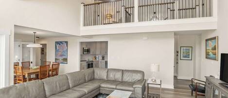 A spacious living area with a large L-shaped sectional sofa, a dining table with chairs, a kitchen in the background, and a balcony with a railing.