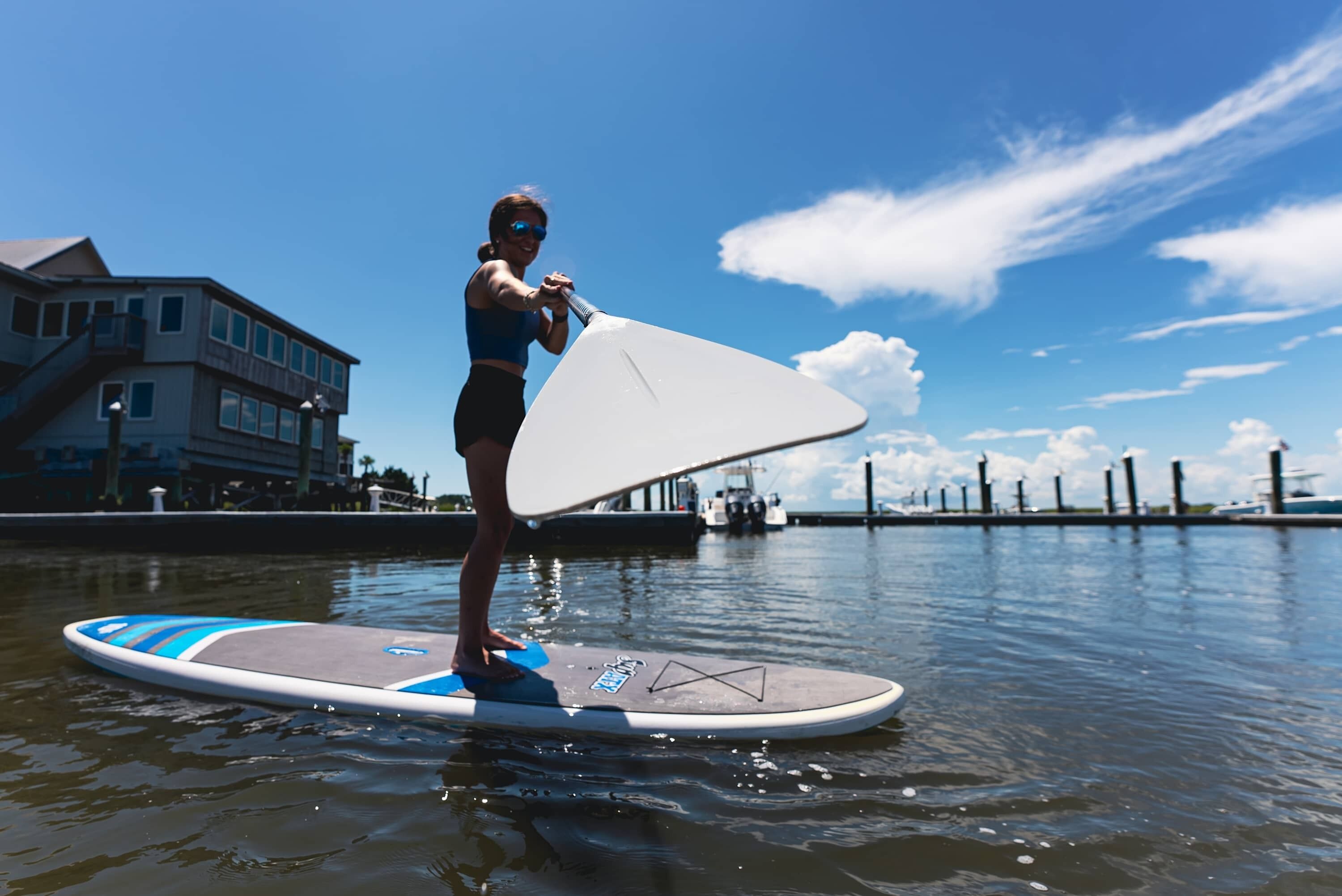 Une personne se tient debout sur une planche de paddle, sur une étendue d’eau, avec des bâtiments en arrière-plan.