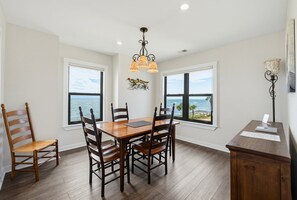Un comedor con una mesa y sillas de madera, ventanas amplias con vistas al océano y una araña.