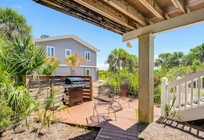 Un patio exterior techado con una parrilla, zona de estar y vistas a una casa de fachada blanca y numerosas ventanas.