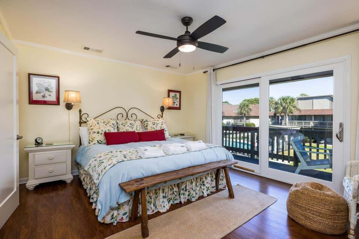 A bedroom with a bed, a nightstand, a bench, a ceiling fan, and a view through a sliding glass door.