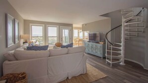 Un salon moderne avec un canapé blanc, un téléviseur, un escalier et de grandes fenêtres.