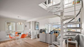 Un salon moderne avec un escalier en colimaçon, un coin repas avec des chaises orange et une grande fenêtre équipée de stores.
