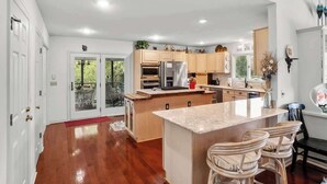 A modern kitchen with wooden cabinets, a central island, and a dining area with wicker chairs. - 395 Tarpon Blvd (Fripp Island)