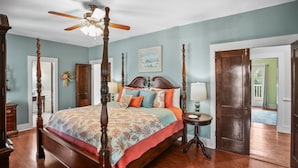 A four-poster bed with a floral bedspread, a ceiling fan, and a view of the outdoors through a door.