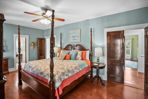 A four-poster bed with a floral bedspread, a ceiling fan, and a view of the outdoors through a door.