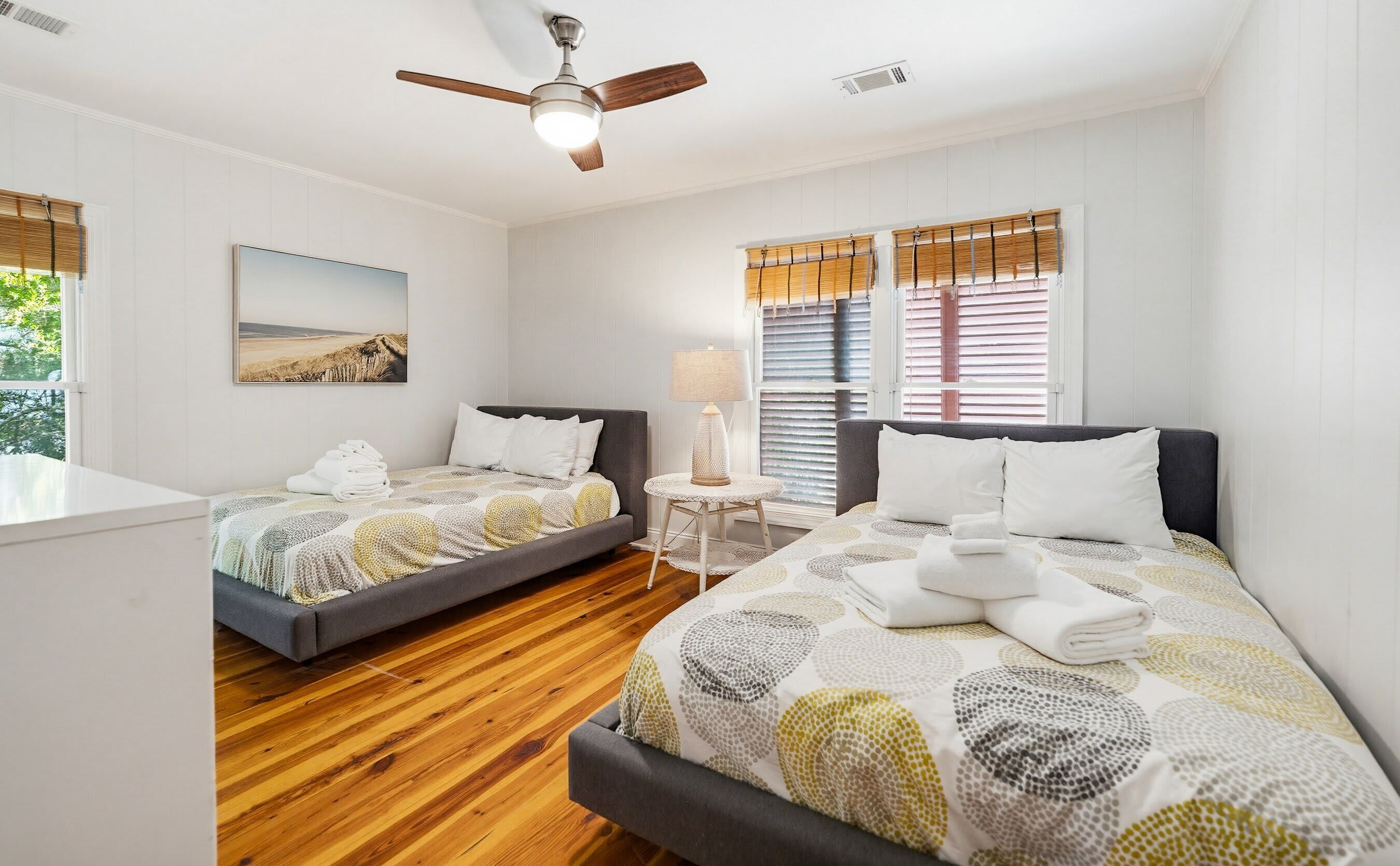 A hotel room with two beds, a ceiling fan, a bedside lamp, and a window with blinds.