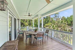 Varanda coberta com mesa e cadeiras de madeira, um ventilador de teto e vista para vegetação tropical.