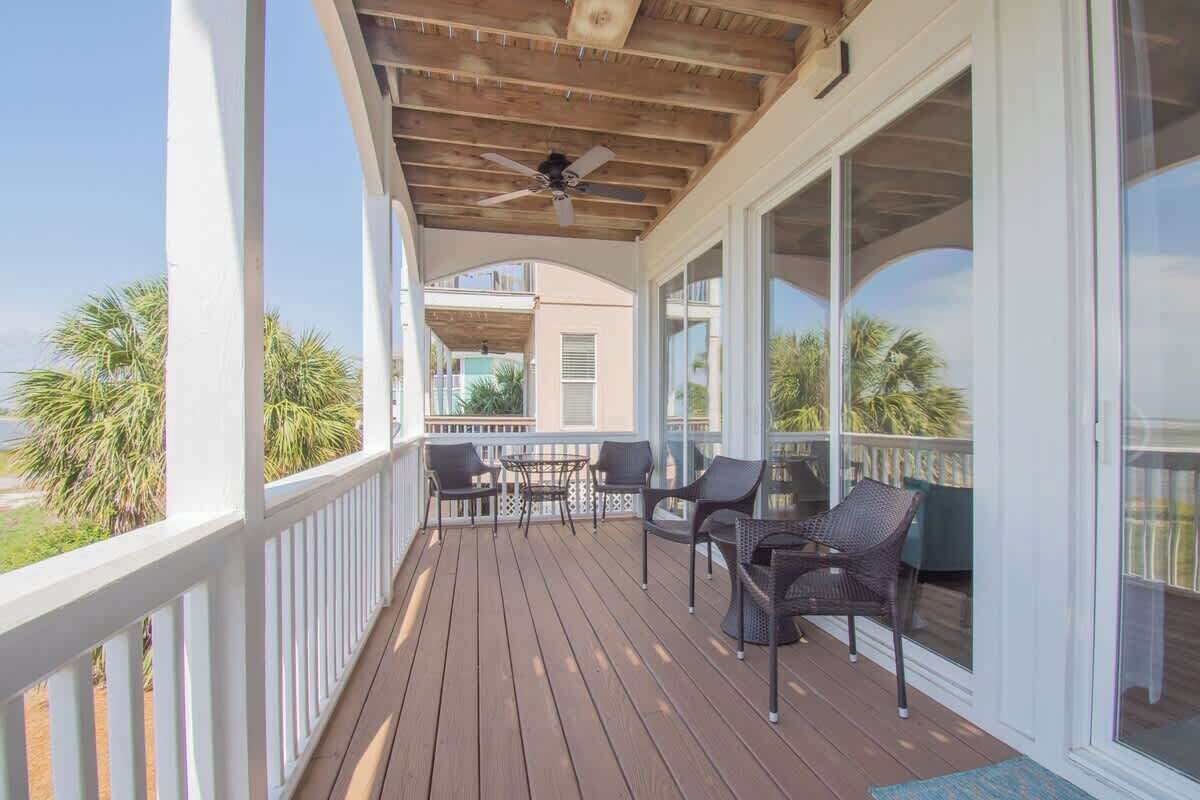 A covered outdoor area with a ceiling fan, a table with chairs, and a view of palm trees and a building in the background.