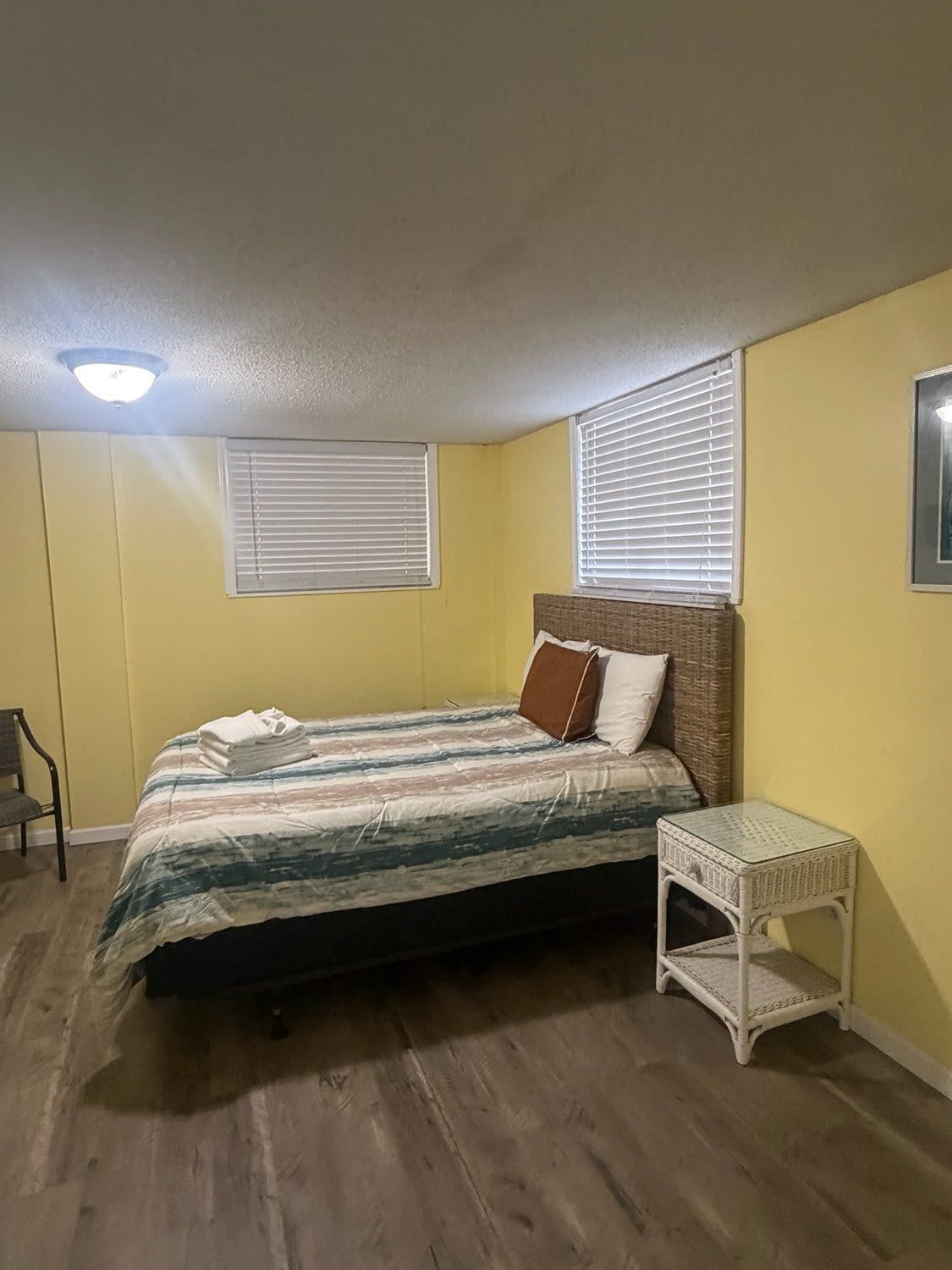 A bedroom with a bed, a chair, a small table, and two windows with blinds.
