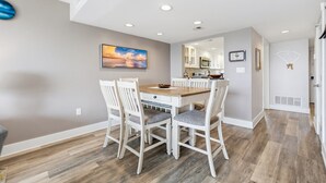 Un coin repas avec une table en bois et des chaises blanches, une cuisine à l’arrière-plan et un grand tableau accroché au mur.