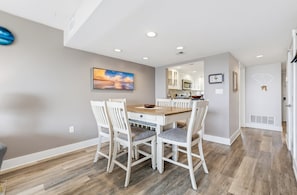 Un coin repas avec une table en bois et des chaises blanches, une cuisine à l’arrière-plan et un grand tableau accroché au mur.