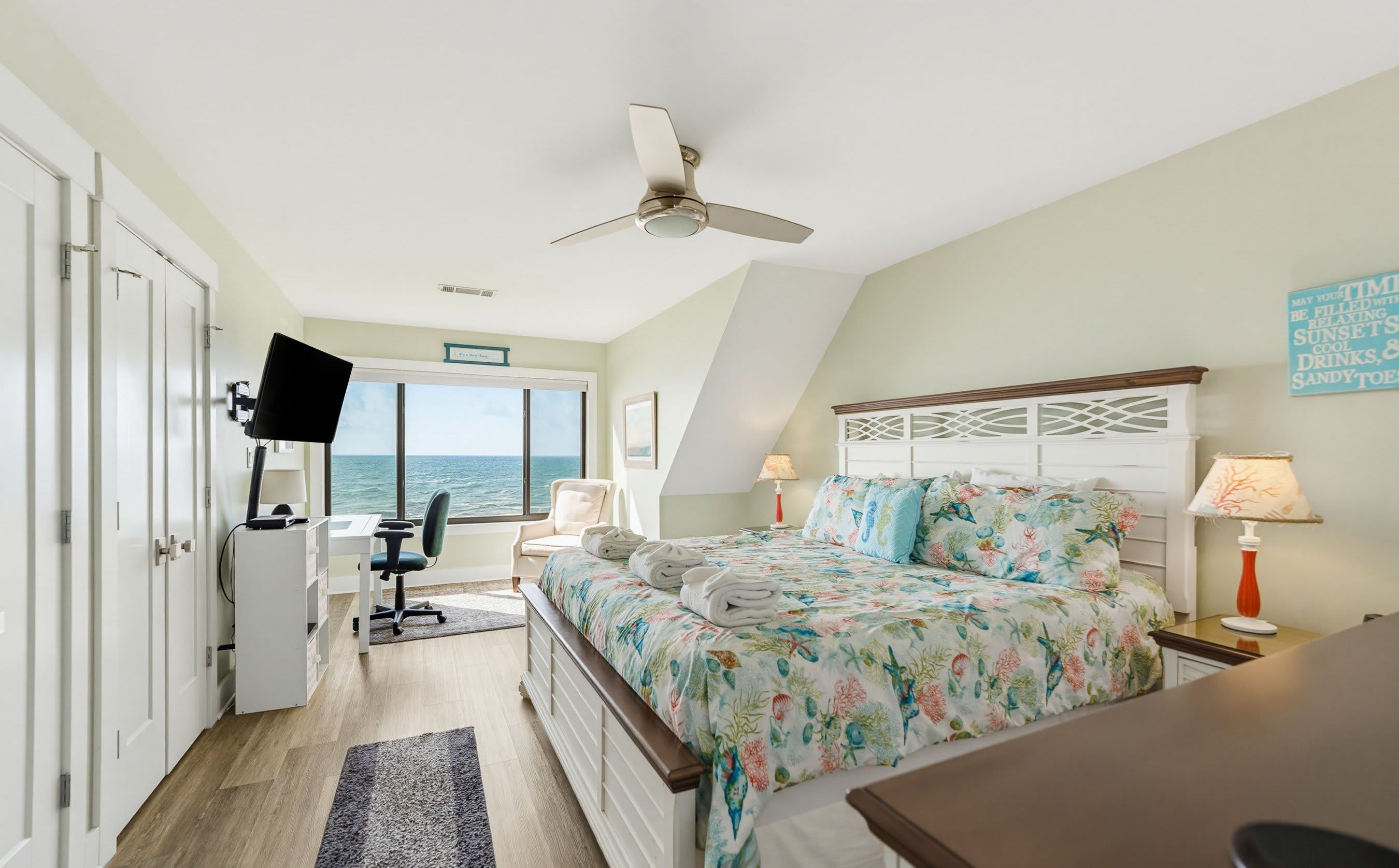 A bedroom with a bed, a desk, a chair, a TV, and a view of the sea.