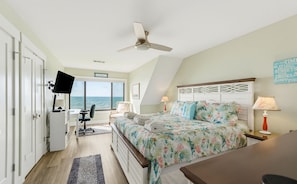 A bedroom with a bed, a desk, a chair, a TV, and a view of the sea.
