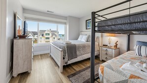Une chambre avec un lit superposé, une table de chevet, une lampe et une vue sur les maisons par la fenêtre.