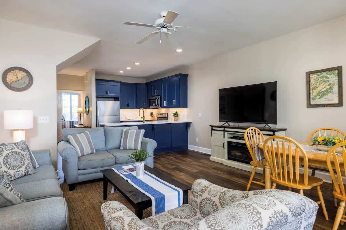 Ein Wohnzimmer mit Sofa, Couchtisch und Fernseher. Es gibt einen Essbereich mit Tisch und Stühlen. Die Küche verfügt über blaue Schränke und eine Mikrowelle.