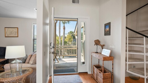 A room with a wooden floor, a wicker table with a glass top, a wicker shelf with a sign, a door leading to a balcony, and a staircase. - 112 Capt John Fripp Villa (Fripp Island)