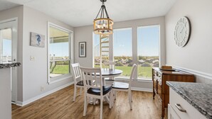 A dining area with a round table and chairs, large windows with blinds, a chandelier, and a clock on the wall.