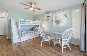 Un lit superposé pour enfants, avec un ventilateur de plafond, une petite table, deux chaises et une fenêtre avec des stores.