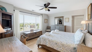 Quarto com cama, escrivaninha, cadeira, cômoda, ventoinha no teto e janela com persianas.