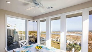 Un coin repas avec une table dressée pour quatre personnes, de grandes fenêtres donnant sur la plage et un ventilateur de plafond.