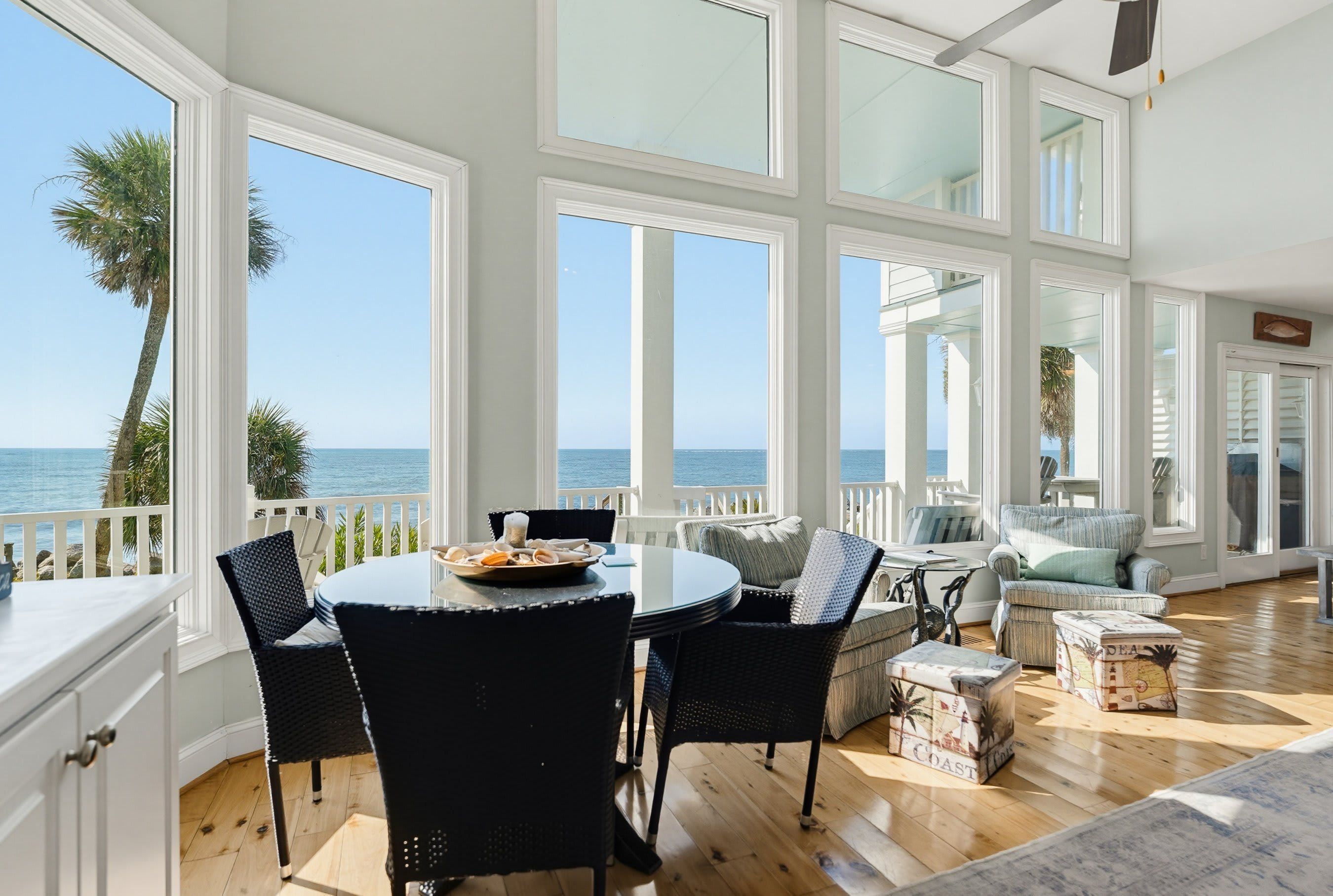 A spacious room with large windows overlooking the ocean, furnished with a round table, chairs, and a sofa.
