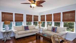 A room with two sofas, a coffee table, a wicker chair, a ceiling fan, and large windows with bamboo blinds. - 266 Tarpon Blvd (Fripp Island)