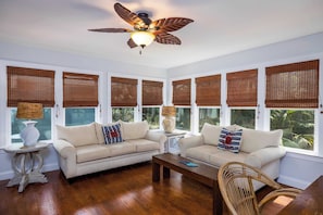 A room with two sofas, a coffee table, a wicker chair, a ceiling fan, and large windows with bamboo blinds.