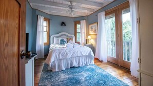 Ein Schlafzimmer mit Bett, Deckenventilator, Fenster mit Vorhängen und Blick auf Grünfläche.