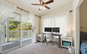 A bedroom with a desk, two monitors, a chair, a ceiling fan, and a balcony with curtains.