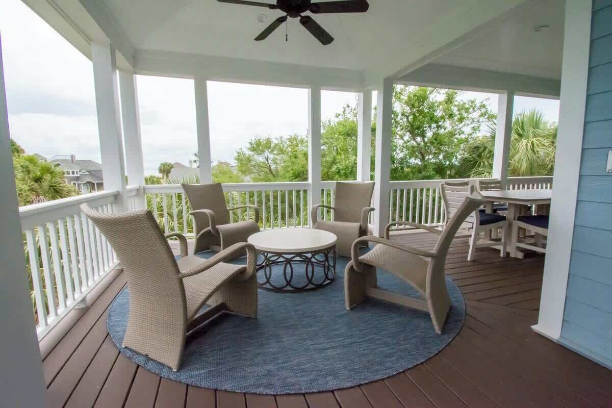 A covered outdoor area with a round table and chairs, a ceiling fan, and a view of greenery and houses.