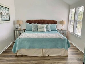 Une chambre à coucher comprenant un lit, deux tables de chevet, une commode, une fenêtre avec stores et une lampe fixée au mur.