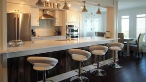 Une cuisine moderne dotée d’un îlot central, de tabourets de bar, d’appareils électroménagers en acier inoxydable et de luminaires suspendus.