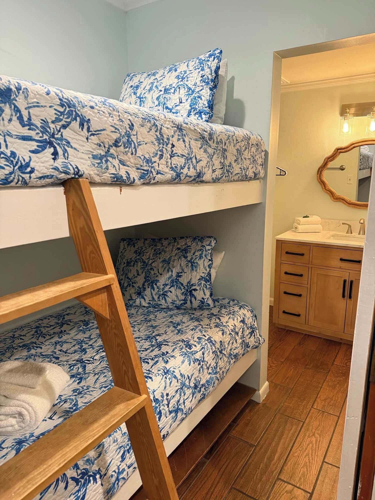 A bunk bed with blue floral bedding, a wooden ladder, a bathroom with a mirror and sink, and wooden flooring.