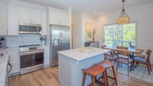 A modern kitchen with white cabinetry, stainless steel appliances, a central island, and a dining area with wooden chairs. - 619 Porpoise Circle (Fripp Island)
