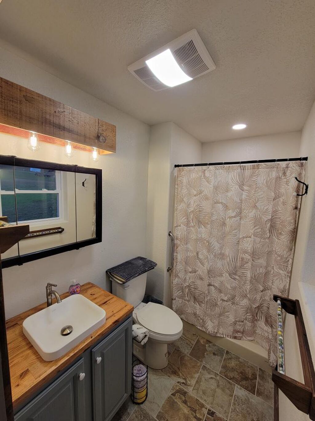 Un baño con tocador de madera, lavabo blanco, inodoro y ducha con cortina estampada.