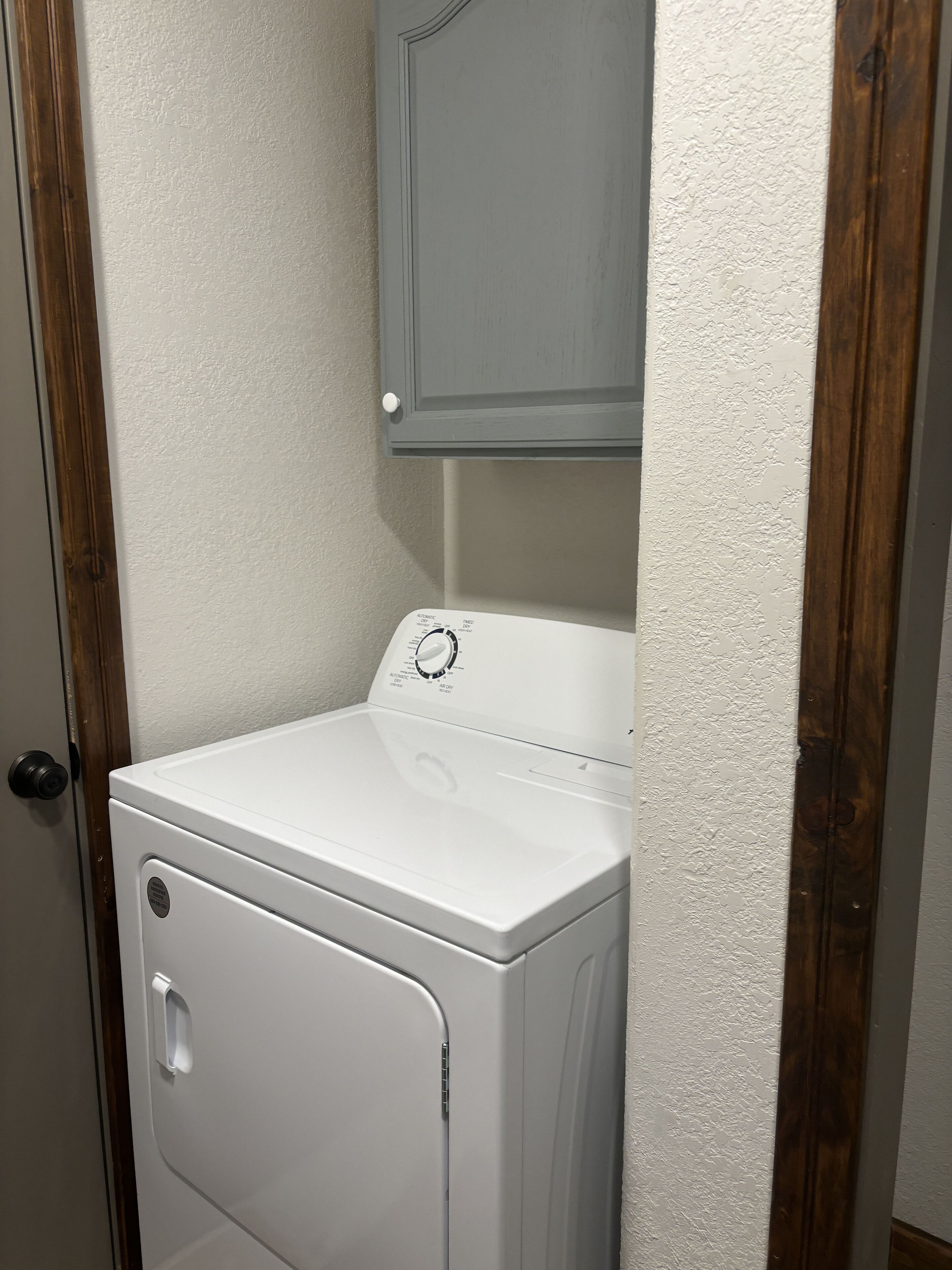 Una lavadora y secadora de carga frontal blancas con panel de control, situadas en un cuarto de lavado con un marco de puerta de madera y un gabinete gris arriba.