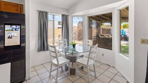Un comedor con mesa de vidrio, sillas blancas, un refrigerador y vista al patio trasero.