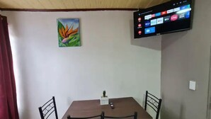 Un coin repas comprenant une table et quatre chaises, un tableau accroché au mur et un téléviseur à écran plat diffusant différentes chaînes.