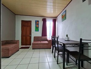 A living room with a sofa, a dining table with chairs, a wooden door, and a window with curtains.