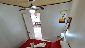 A hotel room with a bed, a ceiling fan, a door leading to a dining area, and a wall with a framed picture.