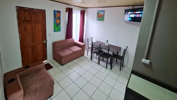 A room with a wooden door, a sofa, a dining table with chairs, and a TV mounted on the wall.