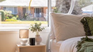 A bedroom with a bed, a bedside table with a lamp, a vase with a plant, and a view of a house through a window.