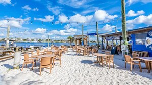 Un café frente a la playa con mesas y sillas de madera, vista al mar y cielo azul con nubes.