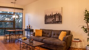 Un salon avec un canapé en cuir marron, une table basse en bois et une table à manger avec des chaises. Une grande fenêtre offre une vue sur les arbres et les bâtiments à l’extérieur.