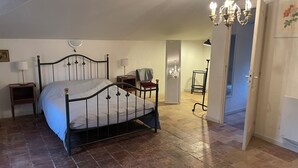 A bedroom with a bed, a chair, a nightstand, a lamp, a chandelier, and a door leading to another room.