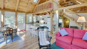 Un intérieur spacieux avec un plafond en bois, un coin repas, une cuisine, un salon doté d’un canapé rose et un escalier menant à un niveau supérieur.