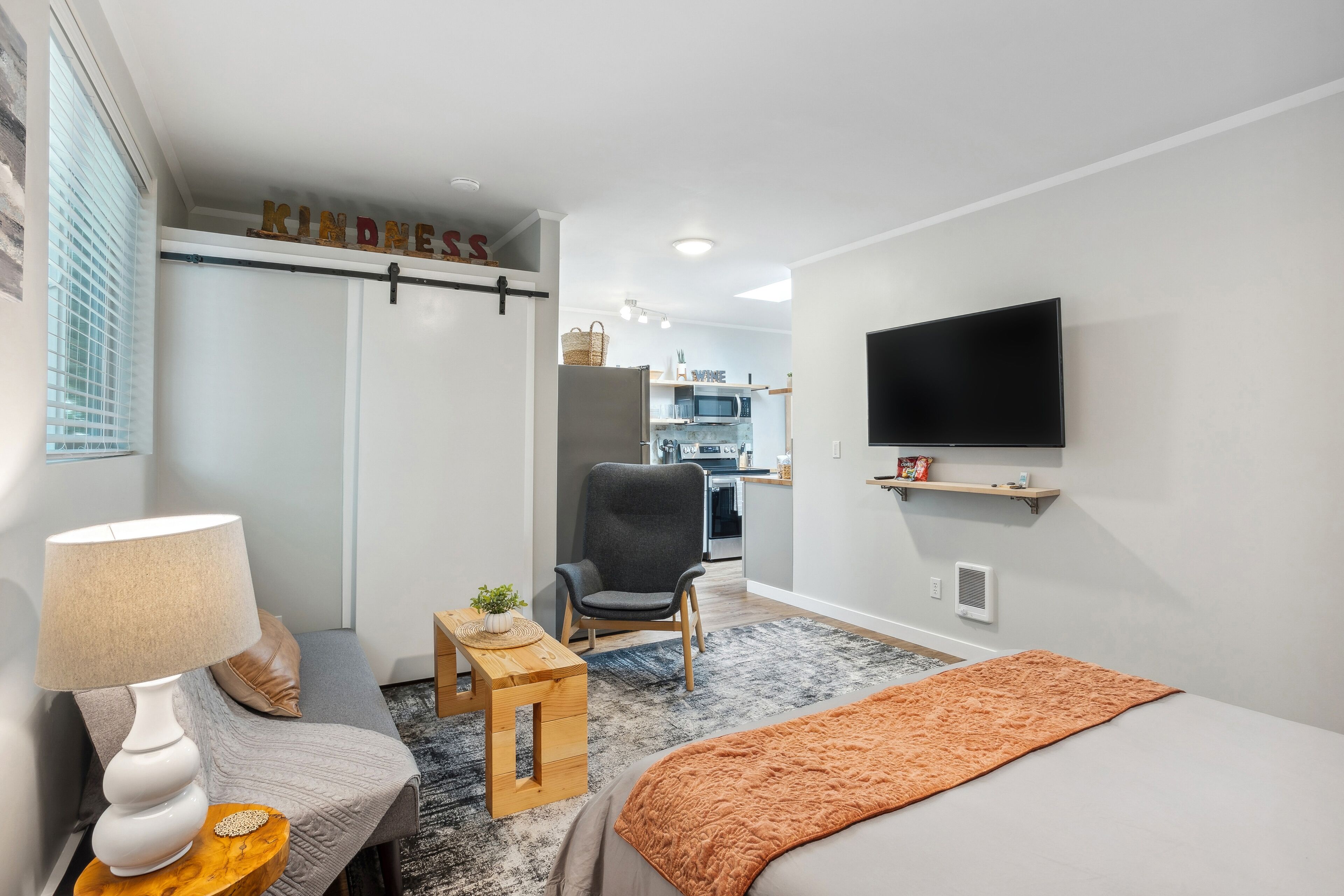 Une chambre d’hôtel moderne avec un lit, un bureau, une chaise, un téléviseur et, au fond, une kitchenette.